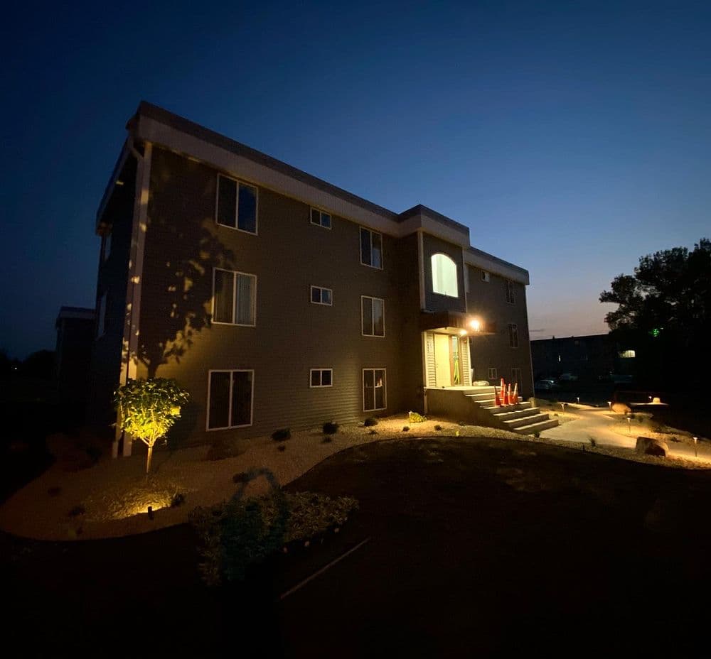 Modern apartment building illuminated at night, surrounded by landscaped garden.