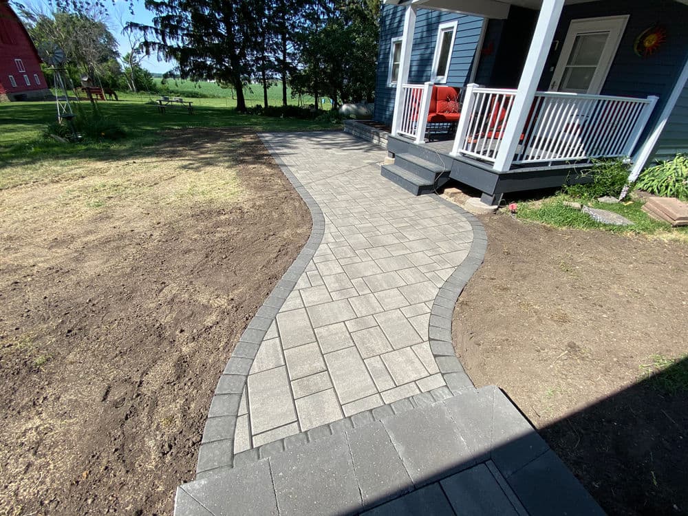 Curved stone pathway leading to a porch in a landscaped yard with trees and a barn.