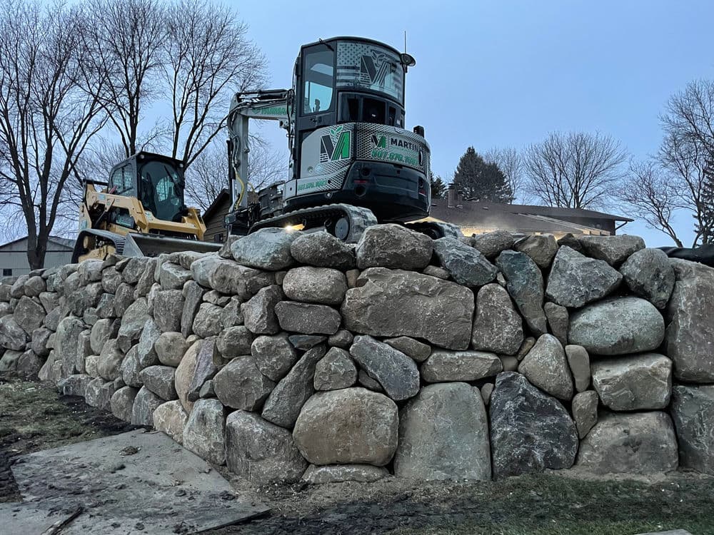 Excavator on top of a stone wall at dusk with trees in the background.