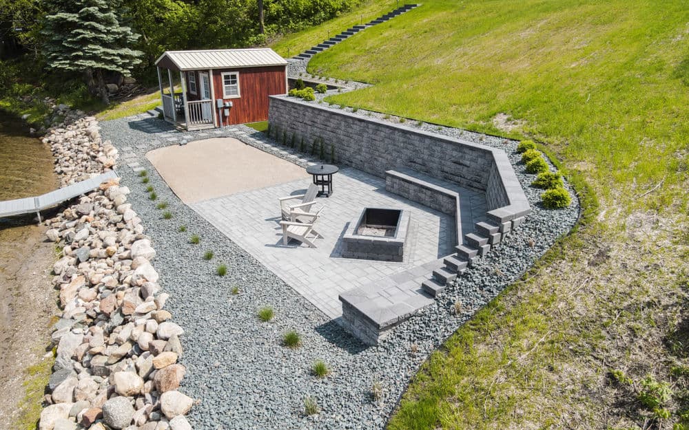 Scenic lakeside patio with fire pit, gray pavers, and cozy shed surrounded by stone landscaping.